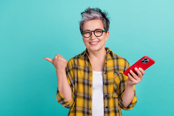 Photo portrait of attractive retired woman point empty space hold device wear trendy plaid yellow clothes isolated on cyan color background