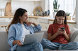 © DimaBerlin - Young smiling mother talking to teen daughter child about being online, modern family using digital devices while resting on sofa at home. Mom chatting speaking with kid about internet safety