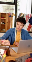 Wall Mural - vertical video of biracial male fashion designer sketching design with laptop in studio, slow motion