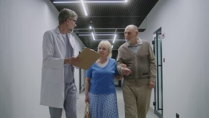 Wall Mural - Elderly couple walks with doctor in clinic corridor. Doctor speaks with patients, discusses medical examination or test results. Medical staff and people in hospital or medical center. Healthcare.