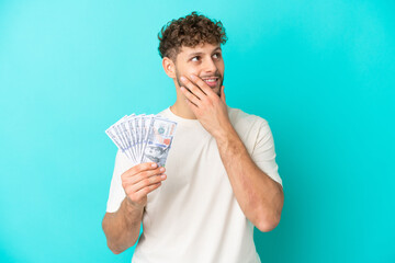 Canvas Print - Young caucasian man taking a lot of money isolated on blue background looking up while smiling