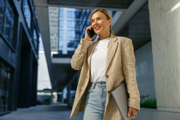 positive female freelancer holding laptop and talking phone standing on modern building background