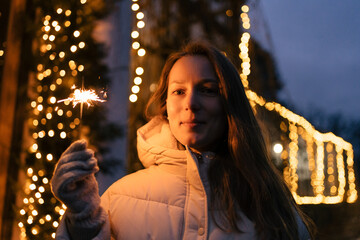 A young woman is holding a bright sparkler and smiling against the background of a garland at night. Concept of New Year and Christmas celebration, coziness, fair and decorated streets.