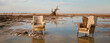 © ADDICTIVE STOCK - Generative AI illustration of two armchairs in the middle of an empty field full of mud and bushes after a flood in blurred background