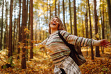 Fototapeta  - Tourist with a hiking backpack, hat walks along a path in the autumn forest. Beautiful woman enjoys a sunny day in nature, feels freedom and breathes fresh air, explores nature.