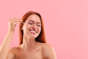 Wall Mural - Beautiful young woman applying cosmetic serum onto her face on pink background, space for text