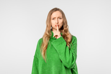 Wall Mural - Serious blonde woman shushing, keep quiet gesture, dressed in green cozy sweater, stand over white background