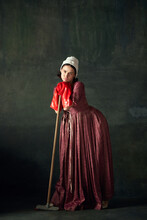Bored And Tired. Beautiful Woman, Medieval Maid In Dress Standing Leaning On Mop In Big Red Rubber Gloves On Vintage Green Background. Concept Of History, Comparison Of Eras, Beauty, Art, Creativity
