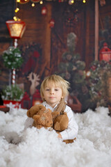 Wall Mural - Cute fashion toddler boy, playing in the snow with teddy bear in front of a wooden cabin log