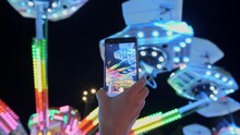 Woman Hands Are Holding Smartphone And Taking Photo Or Shooting Video. Carousel With Lights. Incredible Colorful Flashing Light Of Vintage Carousel.Popular Chair Swing Rides In Amusement Park