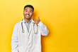 © Asier - Asian doctor in white coat with stethoscope, yellow studio showing a mobile phone call gesture with fingers.