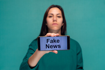 Serious woman holding the mobile phone with the message fake news while looking at camera on isolated on blue