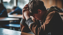 Bullying At High School Concept. Upset Bullied Teen Boy Suffering Sitting Against The Blurred Classroom. Hands Holding His Head, Hiding His Face And Crying. 
