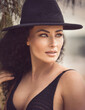 A close-up portrait of a female model. The model has curly hair and poses elegantly in a chic black beach dress and black hat, showcasing her attractive face and physique on a tropical beach.