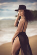Captivating female model with curly hair and a slender sexual body poses elegantly in a chic black beach dress and hat, showcasing her attractive physique at the tropical beach.