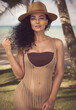 The photo depicts a captivating female model with curly hair and grey eyes, elegantly posed in a beige crochet beach dress and straw hat, against a backdrop of sea and palms