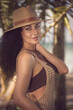 The photo depicts a captivating female model with curly hair and grey eyes, elegantly posed in a beige crochet beach dress and straw hat, against a backdrop of sea and palms