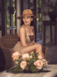 The photo showcases the seductive caucasian female model with curly hair and mesmerising grey eyes. She is dressed in a beige crochet outfit and a hat, sits elegantly in the hall of the luxury hotel. 