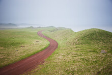 Scenic View Of A Red Dirt Road Surrounded By Tundra With Fog Shrouded Bering Sea In The Background, St. Paul Island, Southwestern Alaska, Summer