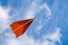 Orangefarbener Fliegender Papierflieger Von Unten Zu Sehen, Im Hintergrund Leicht Bewölkter Blauer Himmel, Rechts Noch Etwas Freier Platz, Horizontal 