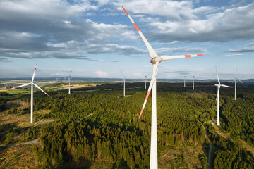  Wind turbines in a hilly forest in front of a partly cloudy, but sunny sky are seen from an aerial view during sunset