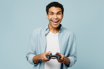 Young smiling cheerful man of African American ethnicity he wears shirt casual clothes hold in hand play pc game with joystick console isolated on plain pastel light blue background studio portrait.