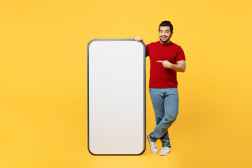 Full body young Indian man wears red t-shirt casual clothes point finger on big huge blank screen mobile cell phone smartphone with area isolated on plain yellow orange background. Lifestyle concept.