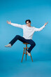 © makistock - Vertical shot of a young cheerful man sitting on a stool isolated on blue background