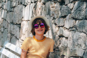 Wall Mural - Beautiful happy girl in straw hat and sunglasses sitting on the bench against stone wall