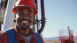 An oilfield worker on an oil rig, wearing a red hard hat, safety glasses, and a gray cover all, with a smile on his face, looking at the camera
