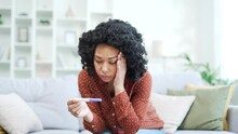 A sad young african american female is holding a pregnancy test with a negative result while sitting on the couch in the living room at home. Disappointed upset black woman dissatisfied and depressed