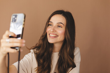 Sticker - Young smiling brunette curly woman wear beige t-shirt doing selfie shot on mobile phone post photo on social network isolated on beige background. Woman make video call, selfie on her mobile phone.