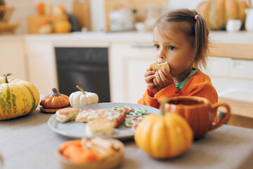 Happy Halloween! Little cute girl eating cookies in the kitchen. Toddler girl in a pumpkin costume eating sandwiches on breakfast. Morning at home.