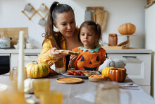 Happy Halloween! Little Cute Girl And Mom Eating In The Kitchen. A Little Girl In A Pumpkin Costume Eats Sandwiches For Breakfast With Her Mother. Morning At Home.