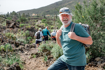 Sticker - Happy caucasian senior man with hat and backpack looks at camera walking the trail on a mountain hike enjoying free time and freedom in nature, healthy lifestyle during retirement