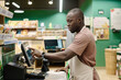 © AnnaStills - African American cashier choosing goods on computer during his work in supermarket