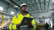 © ihorvsn - African American engineer in hard hat and protective vest is looking through workshop. Man is holding gadget and gesturing at construction site. Employee on background of welding sparks flying.