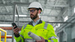 © ihorvsn - Bearded architect inspect ndustrial site. Male holds blueprint and look at phonescreen. Industrial man in reflective vest and hard hat at construction site. Worker at background of work process.