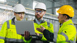 © ihorvsn - Three construction workers in half height look at laptop. Engineers in uniform collaborate with each other in workshop. Man in yellow helmet explains plan of action to employees.
