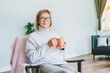 © okrasiuk - Portrait of smiling middle aged woman relaxing with cup of hot drink in scandy style hygge modern interior home. Lady dreaming, enjoy calm mood without stress, well being alone. Cozy autumn
