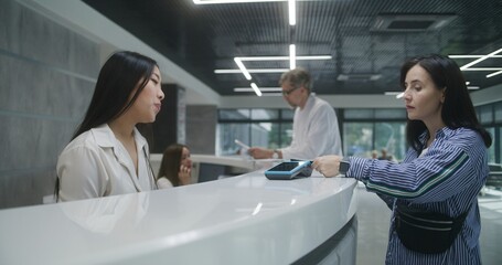 Asian female administrator talks with patient at reception desk in clinic, gives terminal. Woman pays with contactless payment for visit to the doctor using card. Stuff work in modern medical center.