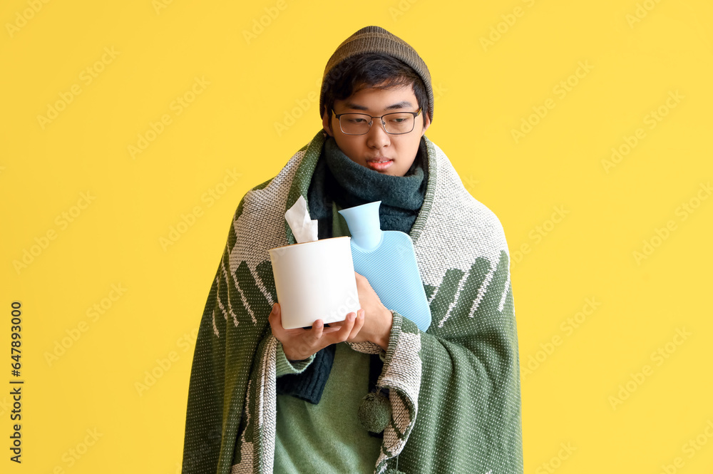 Ill Asian man with hot water bottle and tissue box on yellow background