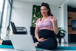 © DusanJelicic - Young pregnant woman in blue and pink sportswear communicates via video call on laptop sitting on yoga mat in living room