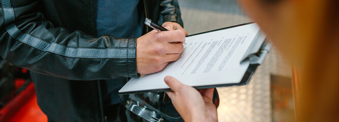 Wall Mural - Unrecognizable biker man holding helmet signing insurance policy to receipt his repaired motorcycle on workshop while female mechanic holding the clipboard