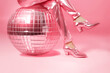 Doll legs in pink shoes and silk trousers with disco ball on pink background.