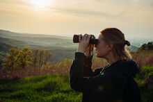 A Hiker Girl Looks Through Binoculars At Nature And Birds Standing On The Top Of A Mountain In The Bright Rays Of Setting Sun Against The Background Of Beautiful Spring Nature And Cloudy Blue Sky