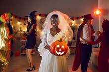 Happy Young Woman In Spooky Costume At Dark Halloween Party With Friends Dressed As Witches, Vampires And Clowns. Smiling Beautiful Black Lady In White Dead Bride Veil Holding Orange Jack-o-lantern