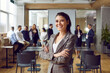 © Studio Romantic - Portrait of successful young business woman looking at camera with confident expression. Female leader stands with her arms crossed against background of people in modern office. Business concept.