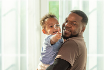 Wall Mural - Happy african black parents dad father throw baby son on piggyback and neck riding teasing fun near window. Black baby son and daddy enjoy teasing kiss cheek and neck riding at window light