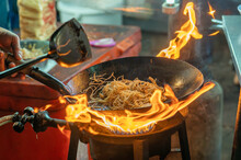 Fried Noodles Cook In Pan With Big Fire Flame Is Hong Kong Style. Pad Thai Favorite And Famous Asian Thai Street Fast Food In Hot Pan, Pad Thai Is Fried Rice Noodle Dish A Street Food Thailand